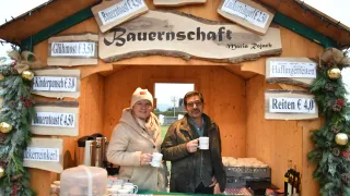 Zwei Personen stehen hinter einer Theke in einer Holzhütte mit ausgewiesenen Preisen an den Schildern. Eine Frau hält eine Tasse und ein Mann hält eine Tasse mit Logo.