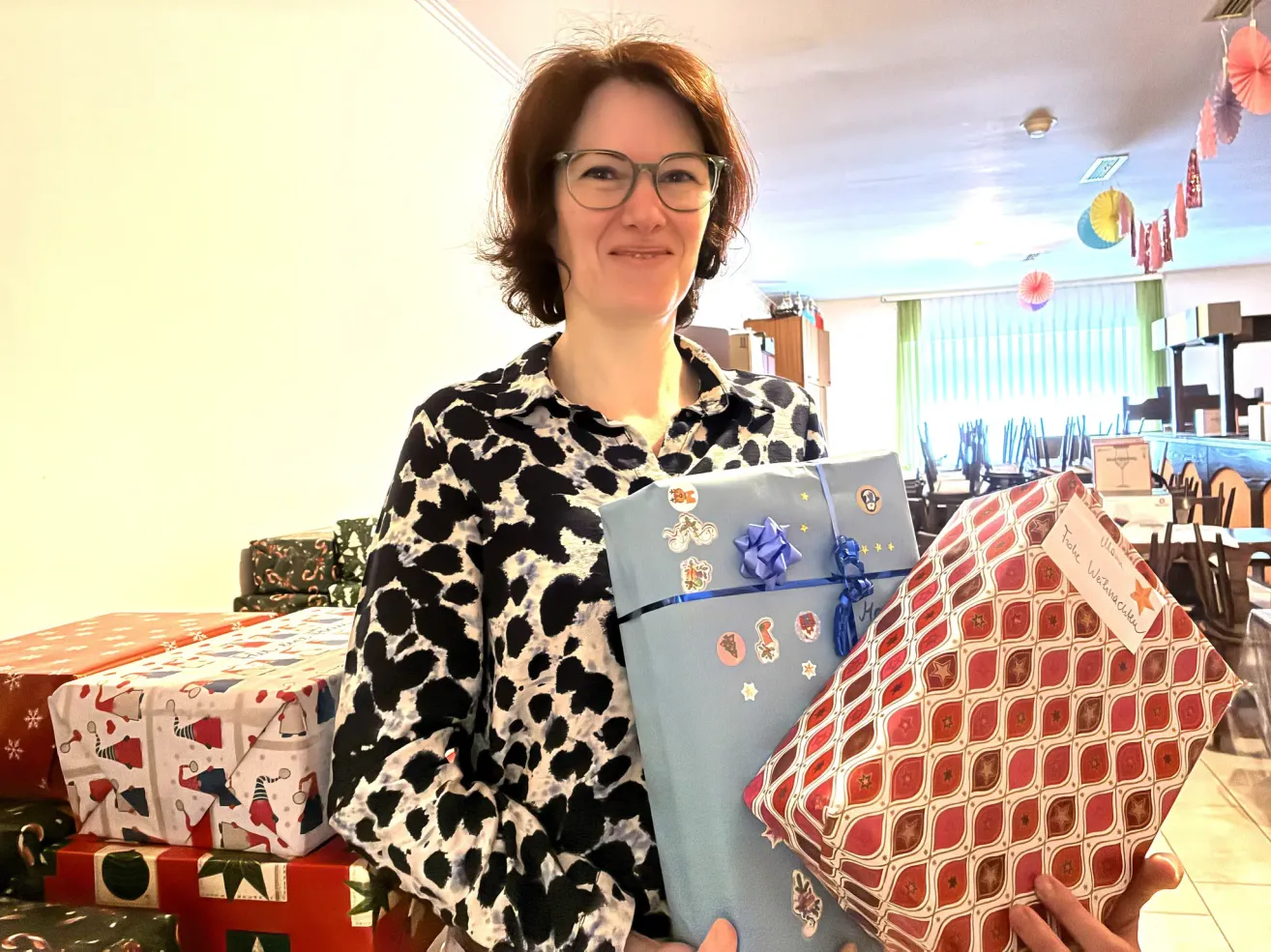 Eine Frau mit Brille hält drei verpackte Geschenke, eines davon hat eine Notiz, die 'Für Walter' sagt. Sie steht vor einem Tisch mit mehreren anderen verpackten Geschenken. Der Raum hat weiße Wände, Deckenleuchten und ein Fenster mit Vorhängen.