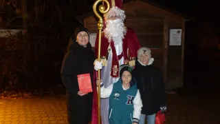 Eine Gruppe von Menschen, darunter eine Frau und zwei Kinder, steht vor einem kleinen Gebäude mit einem Mann, der als St. Nikolaus verkleidet ist. Sie halten alle rote Taschen.