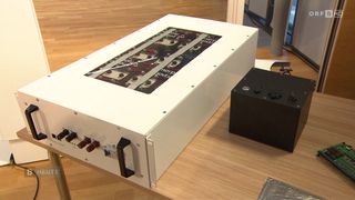 Ein weißer elektrischer Kasten mit einer transparenten Abdeckung, die ein Batteriegitter zeigt, steht auf einer Holzoberfläche. Daneben befindet sich ein kleinerer schwarzer Kasten mit Schaltern.
