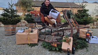 Ein Mann in einer schwarzen Jacke sitzt in einem Schlitten, hält ein Geschenk und ist von verpackten Geschenken und Weihnachtsdekorationen umgeben.