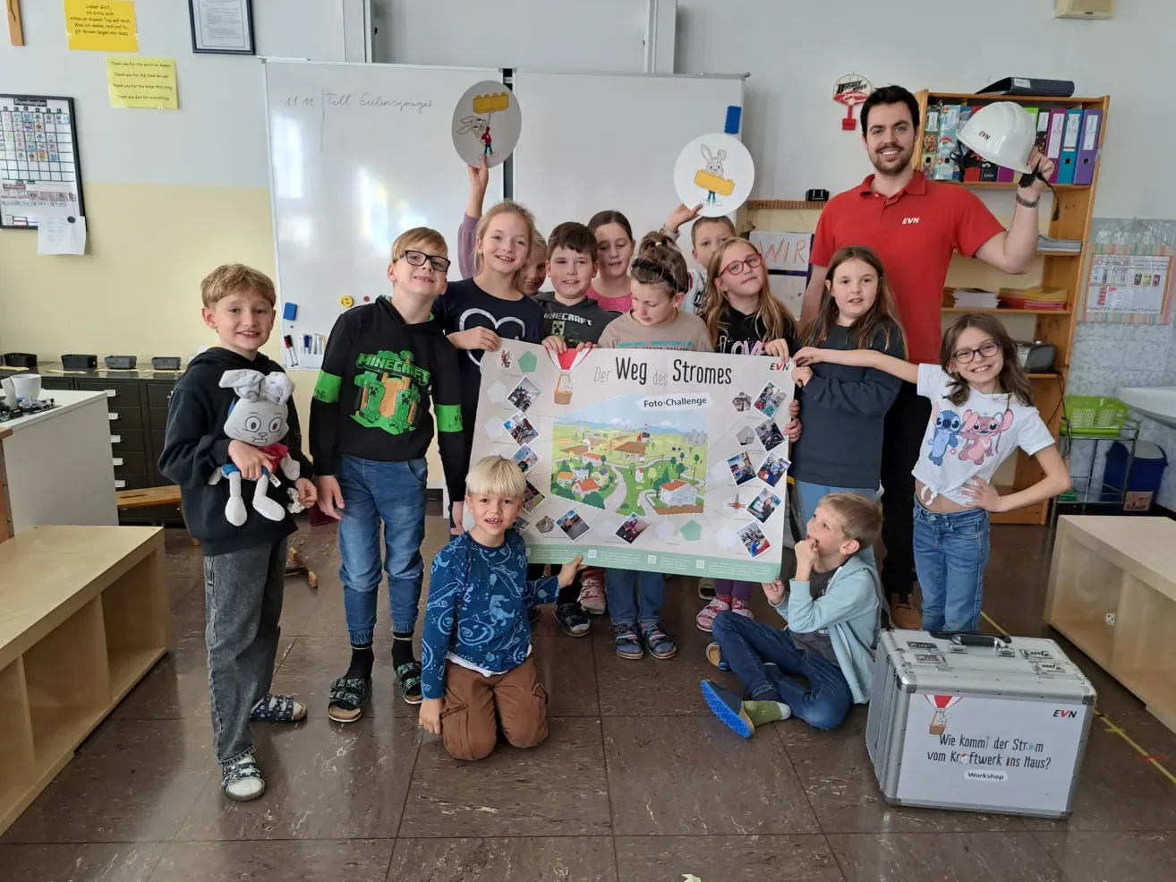 Eine Gruppe von Kindern und ein Mann posiert in einem Klassenzimmer mit einem großen Plakat. Die Kinder halten Bilder hoch. Der Mann steht hinter ihnen.