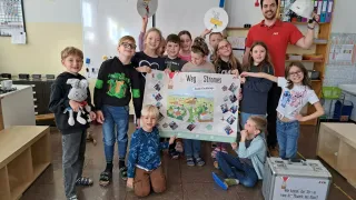 A group of children and a man pose in a classroom with a large poster. The children hold up pictures. The man stands behind them.