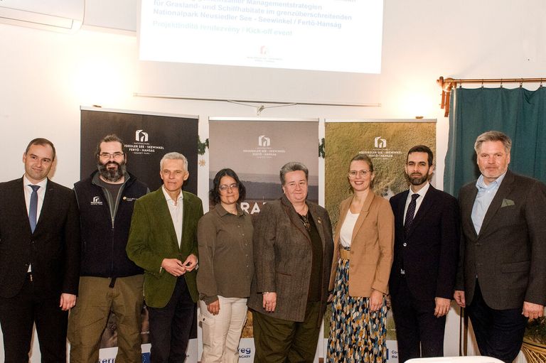 Eine Gruppe von Menschen steht zusammen vor einem Plakat mit einem Bild einer Graslandschaft. Das Plakat trägt den Titel 'Neusiedler See - Seewinkel Fertő-Hanság.' Es sind zwei Männer und drei Frauen, alle formal gekleidet.