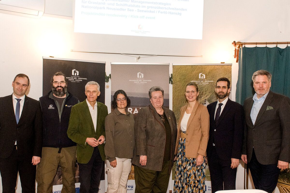 Eine Gruppe von Menschen steht zusammen vor einem Plakat mit einem Bild einer Graslandschaft. Das Plakat trägt den Titel 'Neusiedler See - Seewinkel Fertő-Hanság.' Es sind zwei Männer und drei Frauen, alle formal gekleidet.
