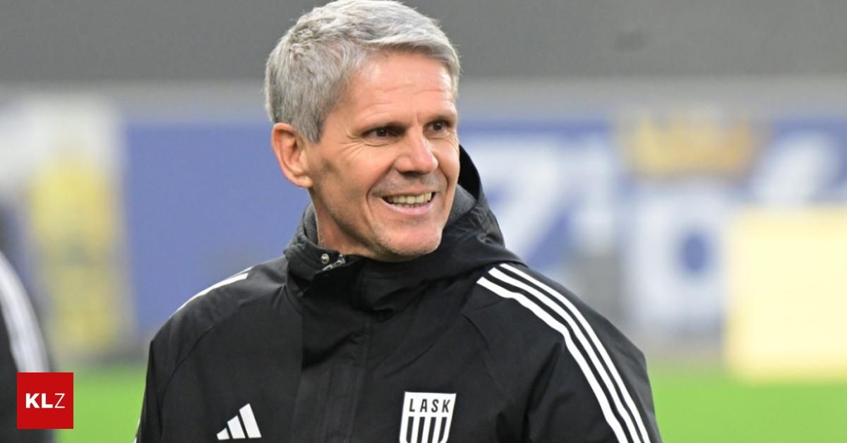 Ein Mann mit einem schwarzen Jacken mit weißen Streifen und dem Wort LASK auf der Vorderseite lächelt.