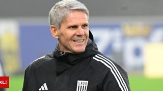 Ein Mann mit einem schwarzen Jacken mit weißen Streifen und dem Wort LASK auf der Vorderseite lächelt.