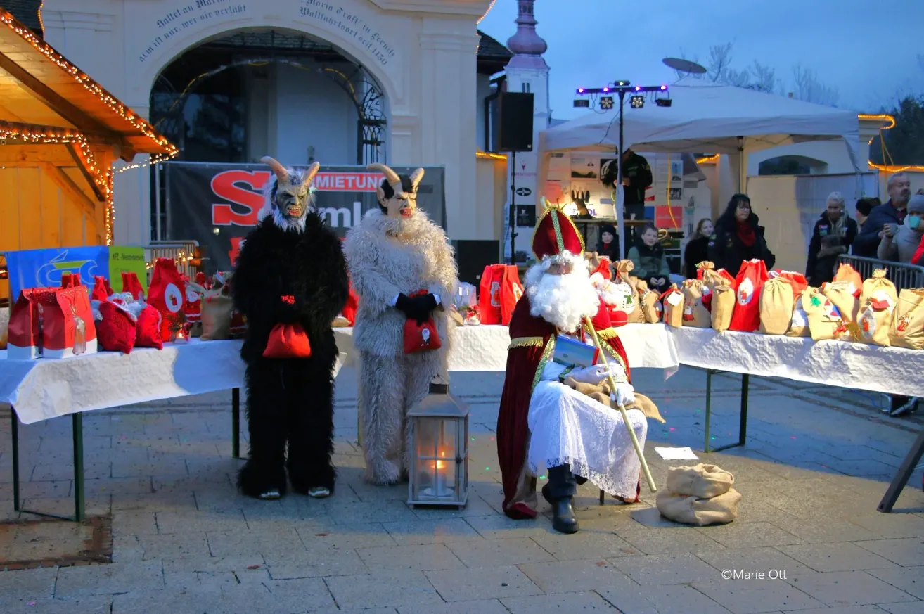 Zwei Personen in Ziegenkostümen stehen neben einer Laterne, während der Weihnachtsmann an einem Tisch mit Geschenken sitzt. Im Hintergrund versammeln sich Menschen.