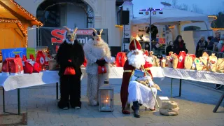 Zwei Personen in Ziegenkostümen stehen neben einer Laterne, während der Weihnachtsmann an einem Tisch mit Geschenken sitzt. Im Hintergrund versammeln sich Menschen.