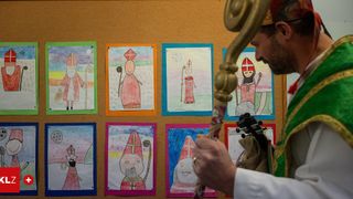 Ein Priester betrachtet Kinderzeichnungen von Heiligen an einer Wand, jede in verschiedenen Farben gerahmt.