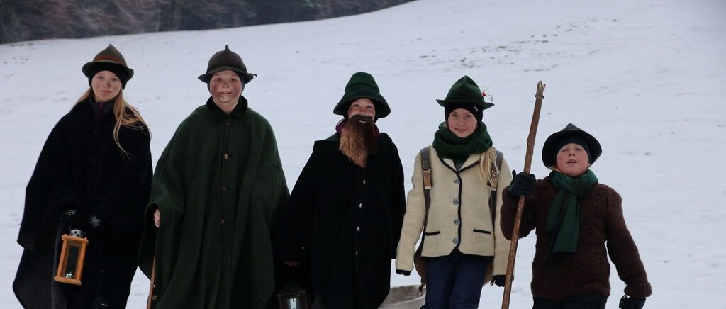 Sechs Personen in Winterkleidung stehen auf einem schneebedeckten Feld. Einer hält einen Holzstab, und andere tragen Rucksäcke. Bäume und Schnee sind im Hintergrund.