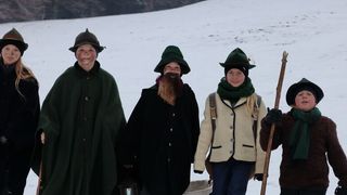 Sechs Personen in Winterkleidung stehen auf einem schneebedeckten Feld. Einer hält einen Holzstab, und andere tragen Rucksäcke. Bäume und Schnee sind im Hintergrund.