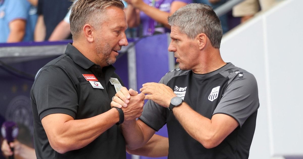 Zwei Männer, wahrscheinlich Fußballtrainer, schütteln sich die Hände in einem Stadion. Einer trägt ein schwarzes Shirt mit Logo, der andere ein graues Shirt mit Logo und einer Uhr.