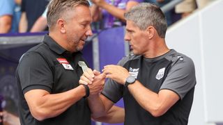 Zwei Männer, wahrscheinlich Fußballtrainer, schütteln sich die Hände in einem Stadion. Einer trägt ein schwarzes Shirt mit Logo, der andere ein graues Shirt mit Logo und einer Uhr.