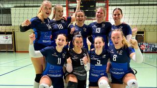 A group of female volleyball players in blue uniforms pose for a photo with smiles, one making a peace sign.