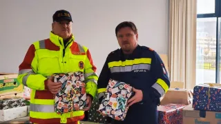 Zwei Männer in Uniform, einer mit einer gelben Jacke und der andere mit einer blauen Jacke, stehen und halten verpackte Geschenke. Sie scheinen Feuerwehrleute zu sein, basierend auf ihren Uniformen und Mützen. Der Raum hat Tische und Stühle, und mehrere Kisten und verpackte Geschenke sind auf den Tischen.