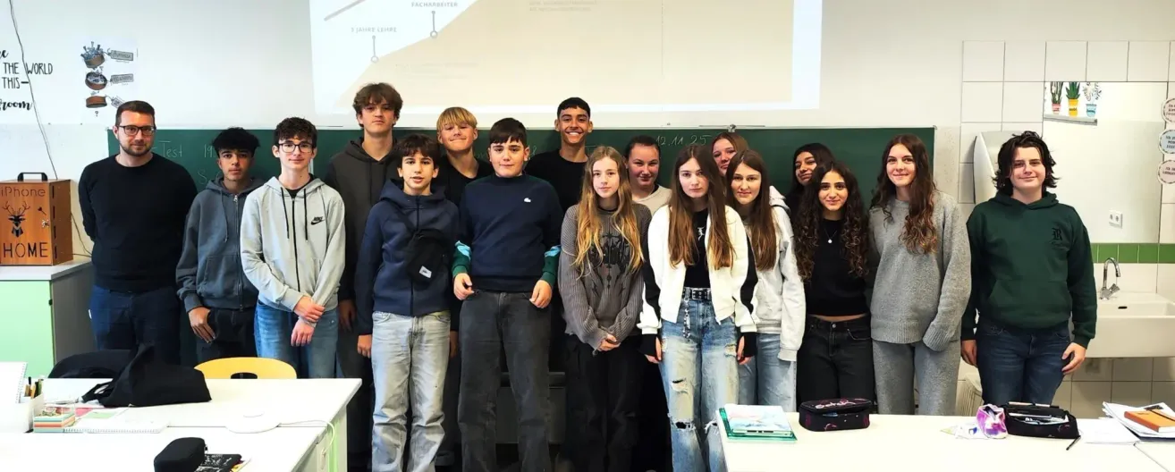 Eine Gruppe junger Schüler steht vor einer Tafel im Klassenzimmer. Sie lächeln und scheinen für ein Foto zu posieren. Ein Schreibtisch ist vor ihnen platziert.