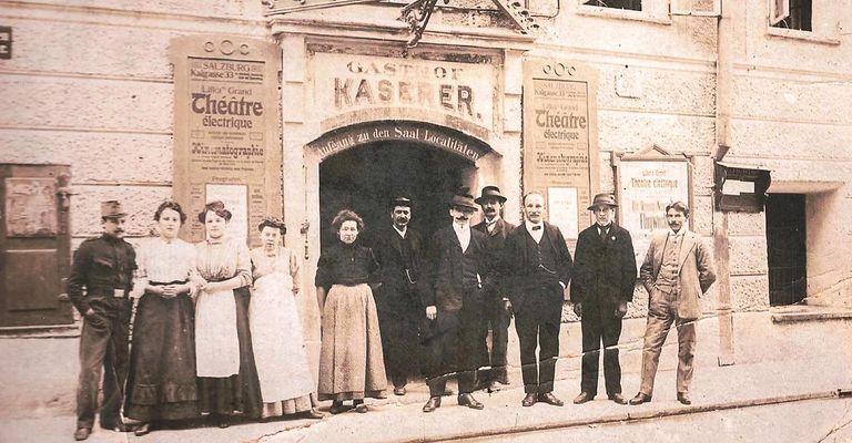 Ein altes Foto einer Gruppe von Menschen, die vor dem Gastof Kaser Gebäude stehen. Sie sind in der Kleidung des frühen 20. Jahrhunderts gekleidet, und es gibt französische und deutsche Schilder am Gebäude.