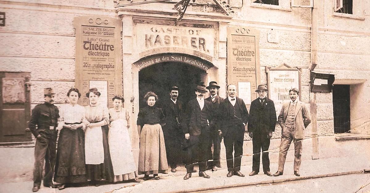 Ein altes Foto einer Gruppe von Menschen, die vor dem Gastof Kaser Gebäude stehen. Sie sind in der Kleidung des frühen 20. Jahrhunderts gekleidet, und es gibt französische und deutsche Schilder am Gebäude.