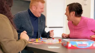 Eine Gruppe von Menschen sitzt um einen Tisch und lächelt und lacht. Sie spielen ein Brettspiel, wobei einer der Männer eine Faust zeigt.