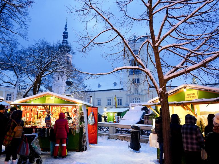 Weihnachtsmarkt in Ljubljana. Eine verschneite Straße mit Leuten, die durch Stände schauen, ein Kirchturm im Hintergrund und ein schneebedeckter Baum.