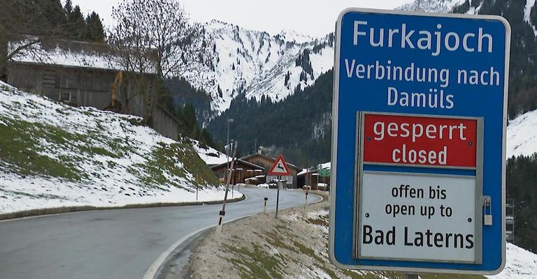 Ein Straßenschild zeigt an, dass der Furka-Pass gesperrt ist. Das Schild ist blau mit weißen Buchstaben. Es zeigt 'gesperrt' in Rot und 'offen b' in Weiß. Schnee bedeckt den Boden und die Berge im Hintergrund.