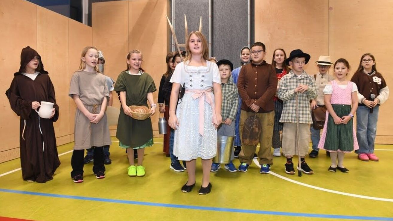 Eine Gruppe von Kindern in verschiedenen Kostümen, möglicherweise für ein Theaterstück, steht auf einem Platz. Eines Mädchens in einem weißen Kleid steht in der Mitte.