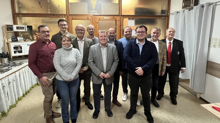 Eine Gruppe von Erwachsenen, darunter Männer und Frauen, stehen zusammen vor einem Gebäude. Sie alle lächeln und scheinen für ein Foto zu posieren.