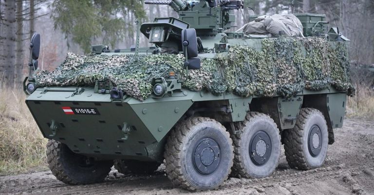 Ein gepanzertes Militärfahrzeug mit Tarnnetz auf einer Schlammstraße. Es hat ein montiertes Maschinengewehr und große Reifen.
