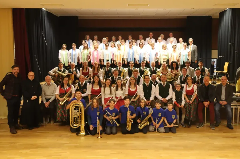 Eine Gruppe von Musikern, darunter Blech- und Holzbläser, posiert für ein Foto auf der Bühne. Sie sind in verschiedenen Kostümen gekleidet und einige halten ihre Instrumente.