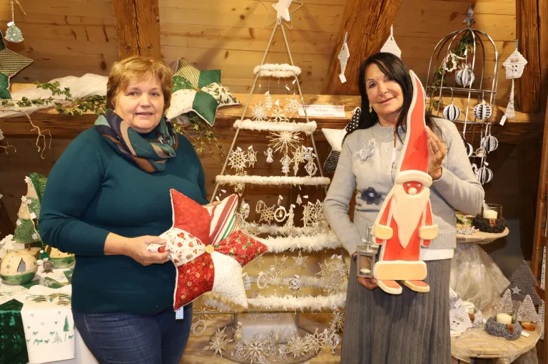 Zwei Frauen stehen in einem Raum mit Holzwänden, eine hält ein Sternkissen und die andere eine Weihnachtsmannfigur. Hinter ihnen steht ein geschmückter Weihnachtsbaum.