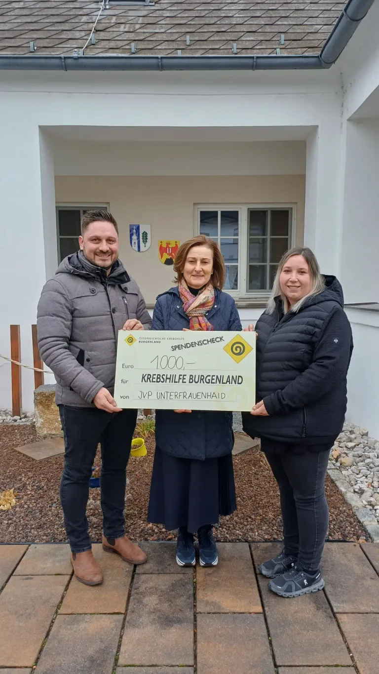 Drei Personen stehen vor einem Gebäude und halten einen Scheck über 1000 Euro mit einem lächelnden Gesicht. Der Scheck ist für Krebshilfe Burgenland. Die Frau in der Mitte trägt einen Schal. Hinter ihnen ist eine Fahne an der Wand angebracht.