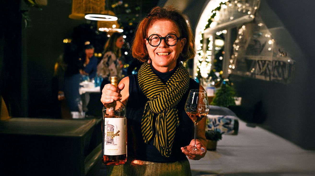 Eine Frau hält eine Flasche Heidi Schrock Wein und ein Weinglas und lächelt für die Kamera in einem festlichen Ambiente.
