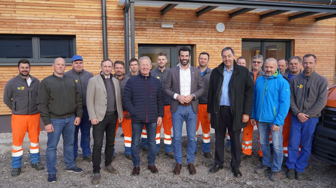 Eine Gruppe von Männern in unterschiedlicher Kleidung, einschließlich Sicherheitsausrüstung, steht vor einem Gebäude. Sie lächeln und scheinen für ein Foto zu posieren.