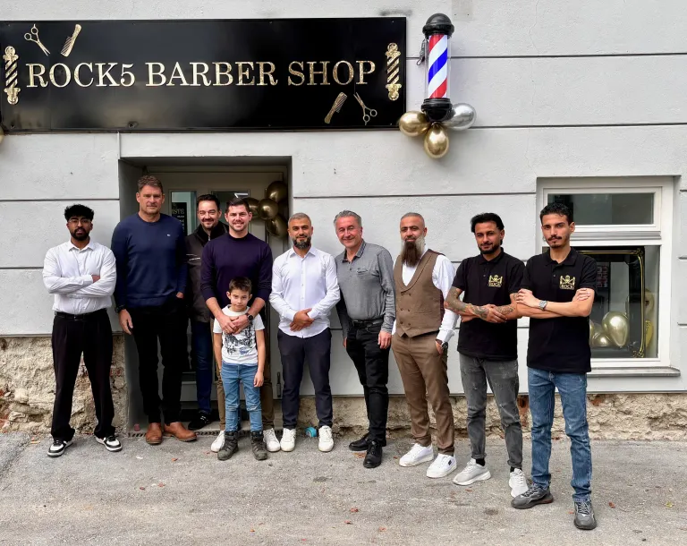 Eine Gruppe von Männern, darunter ein Kind, steht vor einem Friseurladen. Der Laden hat ein schwarzes Schild mit goldener Schrift und einen Friseurpolen draußen. Einige Männer tragen Uhren, und alle sind lässig gekleidet.