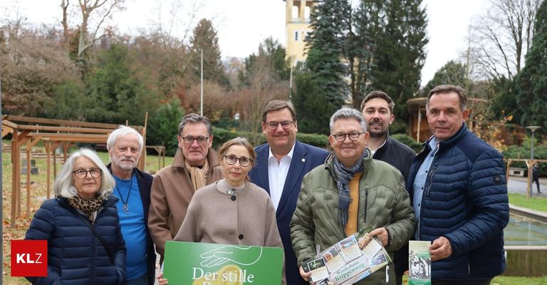 Eine Gruppe von Menschen steht im Freien mit Bäumen und einem Kirchturm im Hintergrund. Die Gruppe besteht aus Männern und Frauen, alle für kühles Wetter gekleidet. Eine Frau hält ein Schild mit der Aufschrift 'Der stille'.