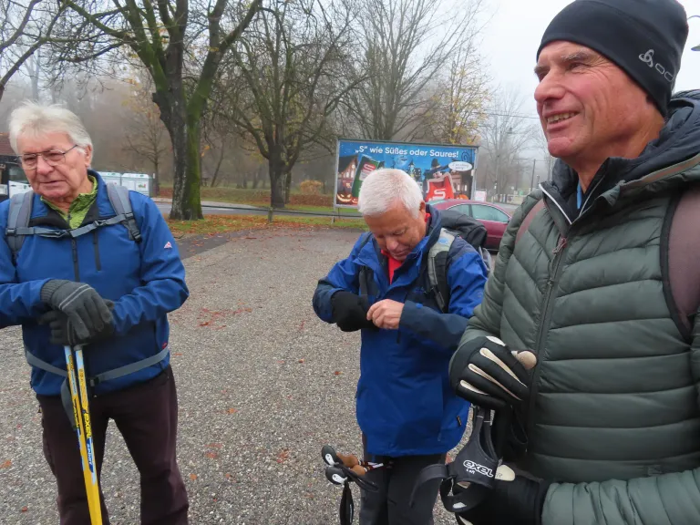 Drei Männer im Freien in einem Park, einer schaut nach unten, während ein anderer Skier hält. Hinter ihnen ein Banner mit einer Kürbisdarstellung.