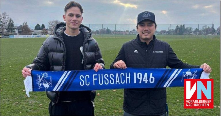Zwei Männer lächeln und halten ein blaues Banner mit der Aufschrift 'SC FUSSACH 1946', stehen auf einem Rasenfeld mit einem Zaun und Bäumen im Hintergrund.