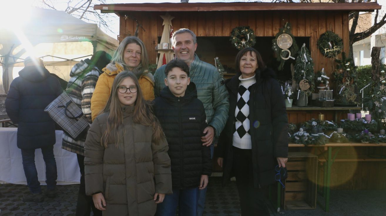 Eine vierköpfige Familie lächelt vor einem hölzernen Stand mit Weihnachtsdekorationen. Der Stand hat Kränze und andere Ornamente. Die beiden Frauen und zwei Jungen sind warm gekleidet.