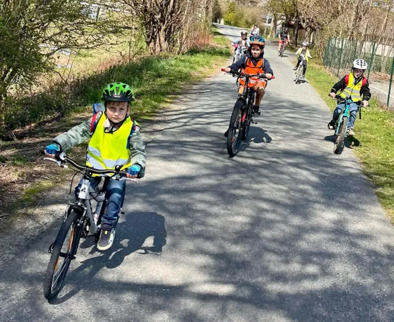 Eine Gruppe von Kindern fährt auf einem Weg in einem Park mit Fahrrädern. Sie tragen Helme und Warnwesten. Der Weg ist von Gras und Bäumen umgeben.
