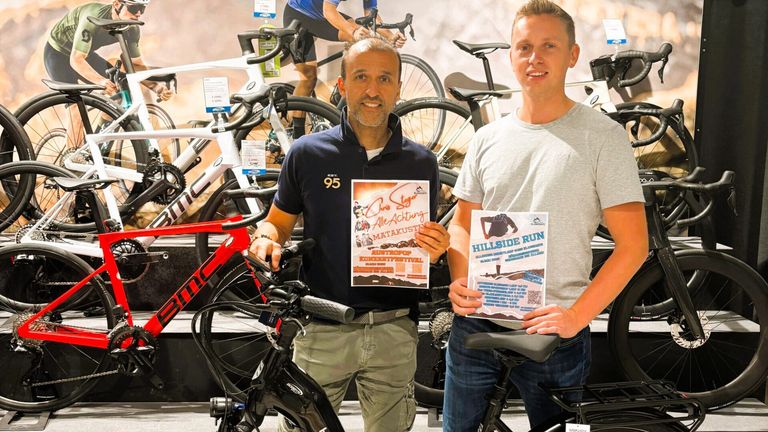 Zwei Männer posieren mit Fahrrädern in einer Ausstellungshalle. Einer hält ein Plakat, der andere eine Broschüre. Fahrräder in verschiedenen Farben sind im Hintergrund zu sehen.