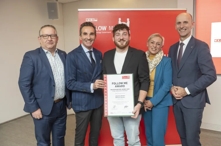 Fünf Personen stehen vor einem roten Hintergrund. Die mittlere Person hält ein Zertifikat mit dem Titel Follow Me Award. Die anderen sind in Geschäftskleidung gekleidet. Die Umgebung wirkt formell, wahrscheinlich eine Preisverleihung.