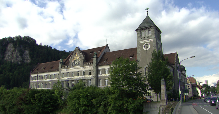 Ein großes, altes Gebäude mit einem Glockenturm steht unter einem bewölkten Himmel, umgeben von Bäumen und Büschen.