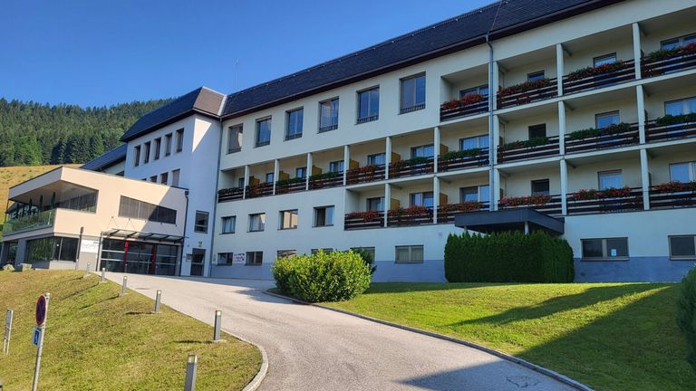 Ein modernes weißes Gebäude mit mehreren Etagen, das Balkone mit roten Blumen aufweist. Die Einfahrt ist mit Sträuchern gesäumt und führt zum Eingang.