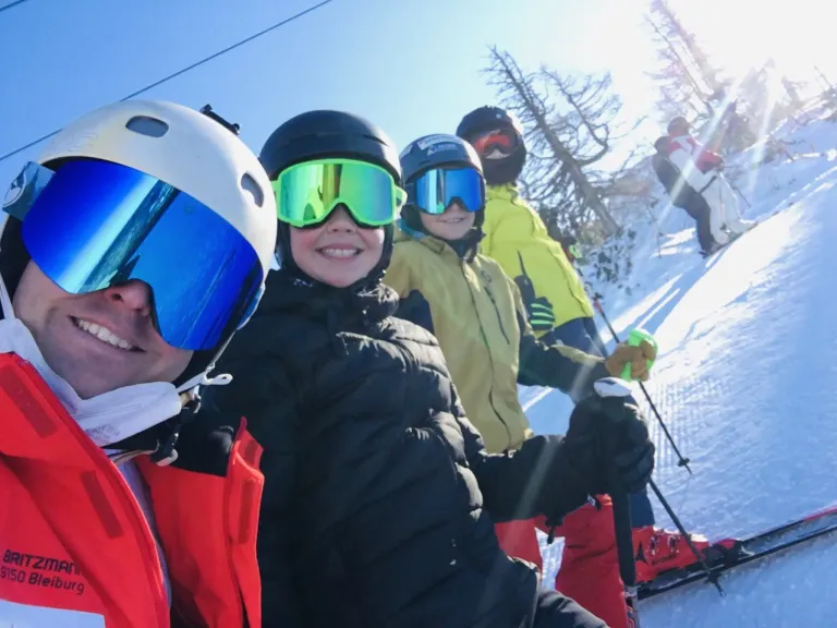 Vier Personen stehen gemeinsam auf einem verschneiten Hang und tragen Winterausrüstung und Helme. Im Hintergrund sind Bäume zu sehen.