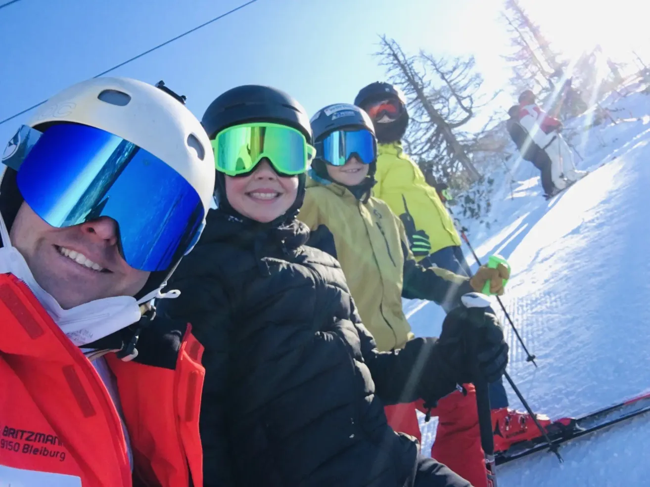 Vier Personen stehen gemeinsam auf einem verschneiten Hang und tragen Winterausrüstung und Helme. Im Hintergrund sind Bäume zu sehen.