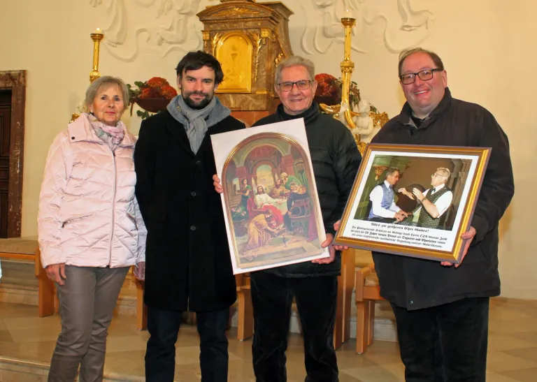 Vier Personen stehen in einer Kirche, lächeln und halten gerahmte Bilder. Eines ist ein religiöses Gemälde, die anderen sind Porträts. Dahinter ein Altar mit Kerzen und einer Statue.