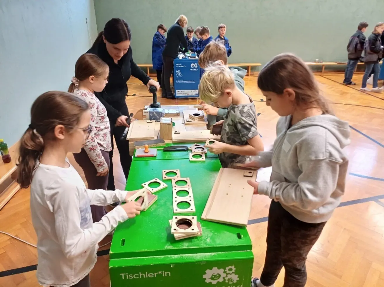 Mehrere Kinder, darunter Mädchen und Jungen, nehmen an einem praktischen Workshop in einem Raum mit einem grünen Tisch und verschiedenen Holzobjekten teil. Eine erwachsene Frau beaufsichtigt die Aktivität.