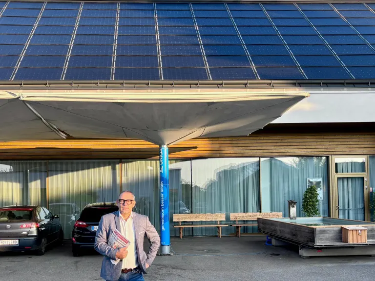 Ein Mann steht vor einem modernen Gebäude mit Solarpanelen und hält ein Buch. Hinter ihm sind Bänke in der Nähe eines Brunnens platziert. Autos sind vor dem Gebäude geparkt.