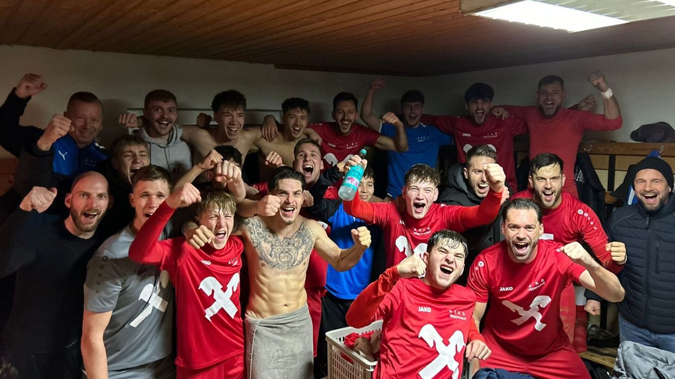 Eine Gruppe von Fußballspielern in roten Trikots mit 'X' darauf feiert in einer Umkleidekabine. Sie jubeln und zeigen ihre Freude.